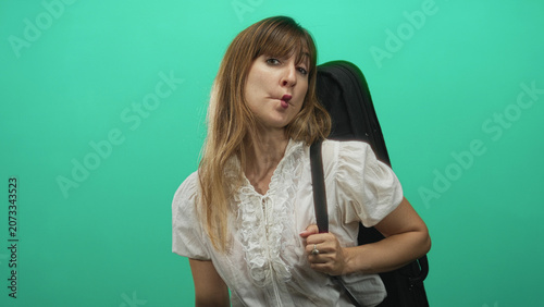 Foto Young woman holding guitar case strap and making puckered lips in a green studio setting; playful travel
