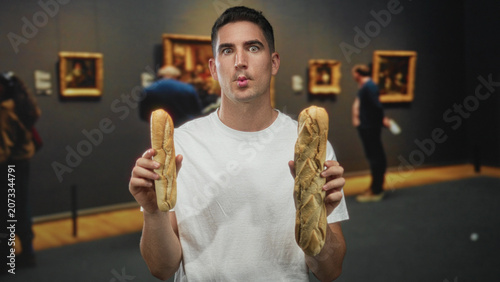 Billede på lærred Man holding two baguettes in hands inside art gallery building, puckering lips while facing camera; playful mischief