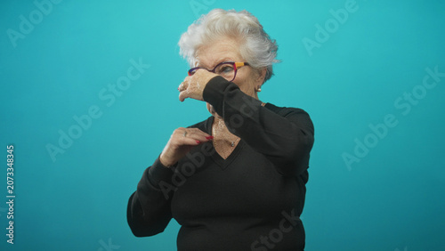 Foto Elderly woman in black sweater pinches her nose with hand and closes eyes in turquoise studio beside glasses; disgust aversion