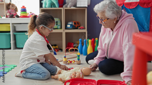 Photos Woman kneeling with girl roleplaying doctor, holding a toy stethoscope and examining a baby doll in a daycare playroom; intergenerational bonding tenderness