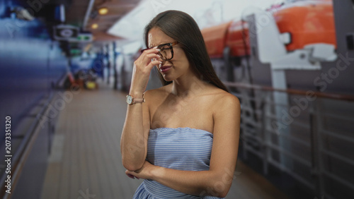 Fotografie Woman pinches bridge of nose with hand on cruise ship deck, glasses and strapless dress visible; frustration travel delay
