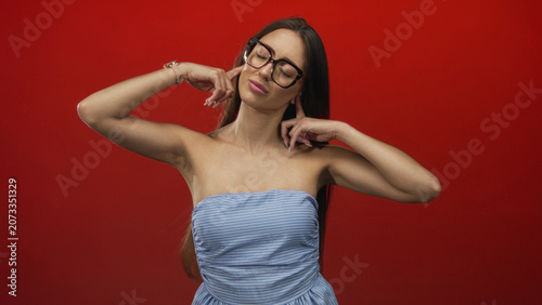 Canvastavla Woman in strapless dress and glasses pressing fingers to her ears, bare shoulders visible in red studio; annoyance defiance