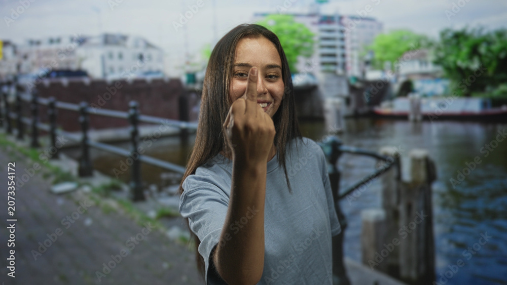 Fototapeta premium Woman showing middle finger directly to camera on a canal street in amsterdam wearing a light blue tshirt by a railing and boat; raw defiance.
