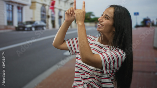 Photos Young woman points fingers upward on city street while smiling and looking up, wearing striped shirt and long hair; joy playful energy