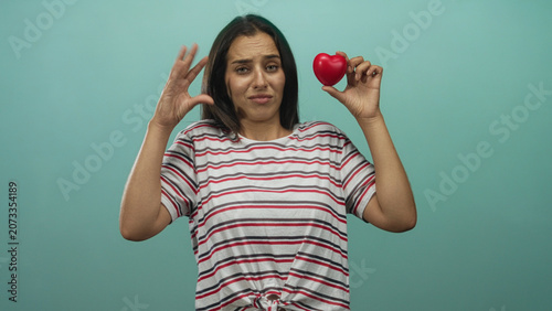 Foto Young hispanic woman holds red heart with raised hand gesture in studio against turquoise backdrop; skepticism