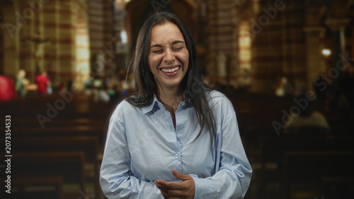Photos Woman laughing with face toward camera, hands clasped over stomach inside a catholic church nave with wooden pews and stone arches; joy faith