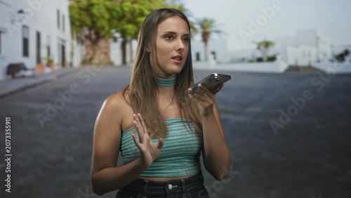Fotografie Young woman holding smartphone with hand raised in a speaking gesture on a street near white buildings; concern communication