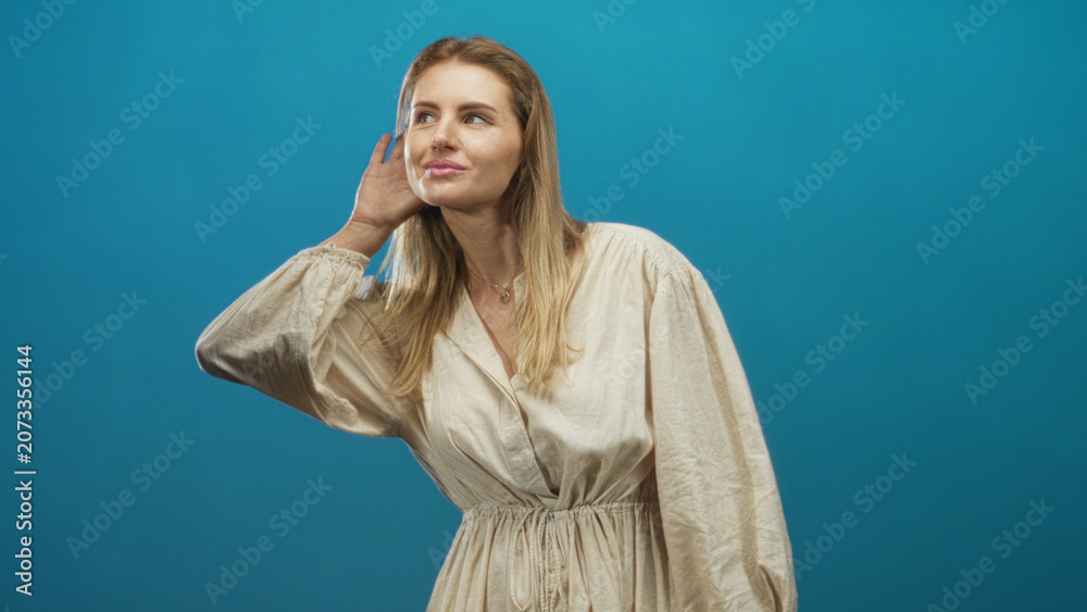 Obraz premium Woman cupping hand to ear in studio against blue backdrop wearing beige dress and necklace; curiosity attentiveness.