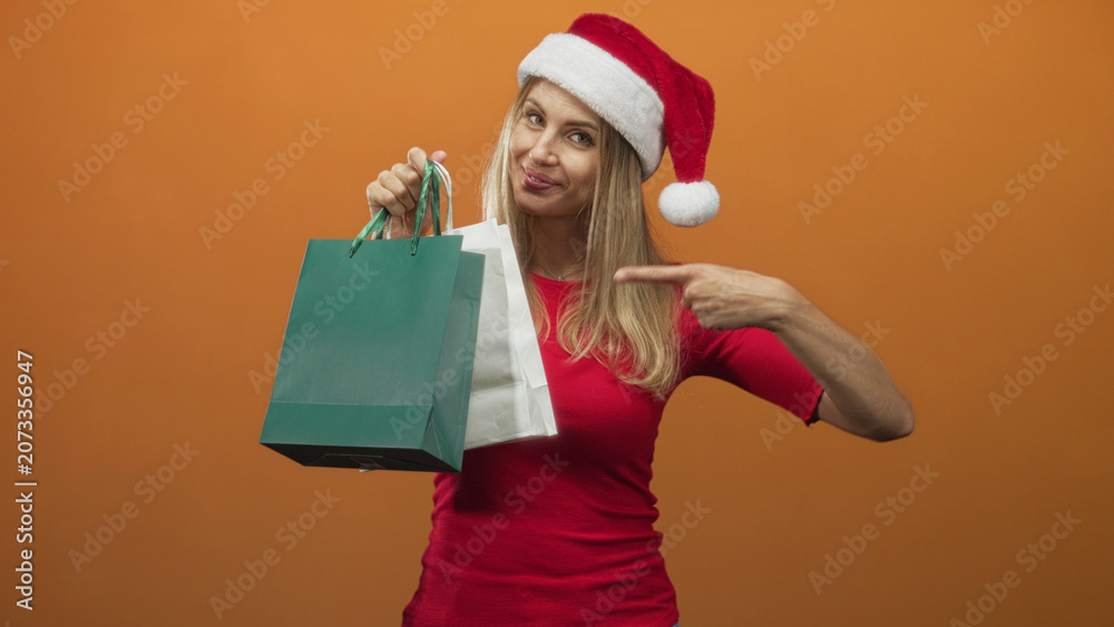 Fototapeta premium Blonde woman in santa hat holding green and white shopping bags points finger to bags in studio; holiday cheer.
