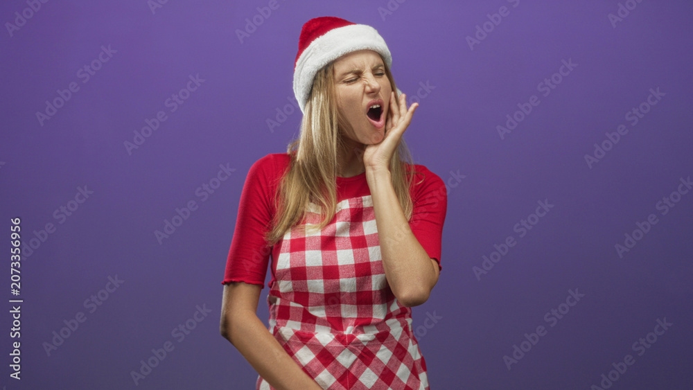 Fototapeta premium Woman with hand on cheek wincing and touching jaw in a studio wearing santa hat and red checked apron; holiday pain.
