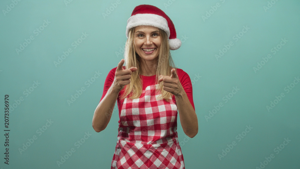 Fototapeta premium Woman pointing fingers at camera, smiling, wearing red checked apron and santa hat in studio with teal backdrop and soft lighting; holiday joy.