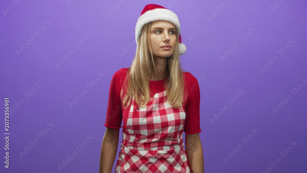 Fototapeta premium Woman wearing santa hat and red checked apron stands centered and looks to side with bare forearms visible against purple backdrop in studio; holiday boredom.