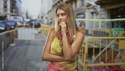 Schilderij op canvas Woman biting fingernail with crossed arms beside street construction barrier, hand at mouth showing nervous gesture; anxiety uncertainty waiting