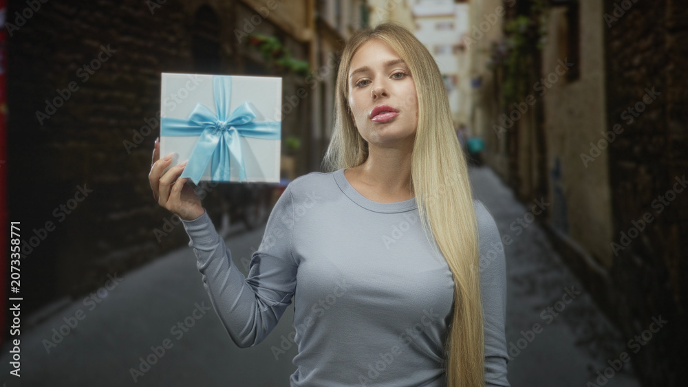 Fototapeta premium Woman holding gift box with blue ribbon, hand raised presenting, long blonde hair in narrow cobblestone street; gift giving anticipation.