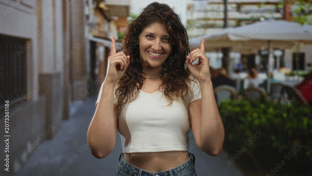 Fototapeta premium Woman points both index fingers upward on a street terrace, smiling with exposed midriff visible; joy.