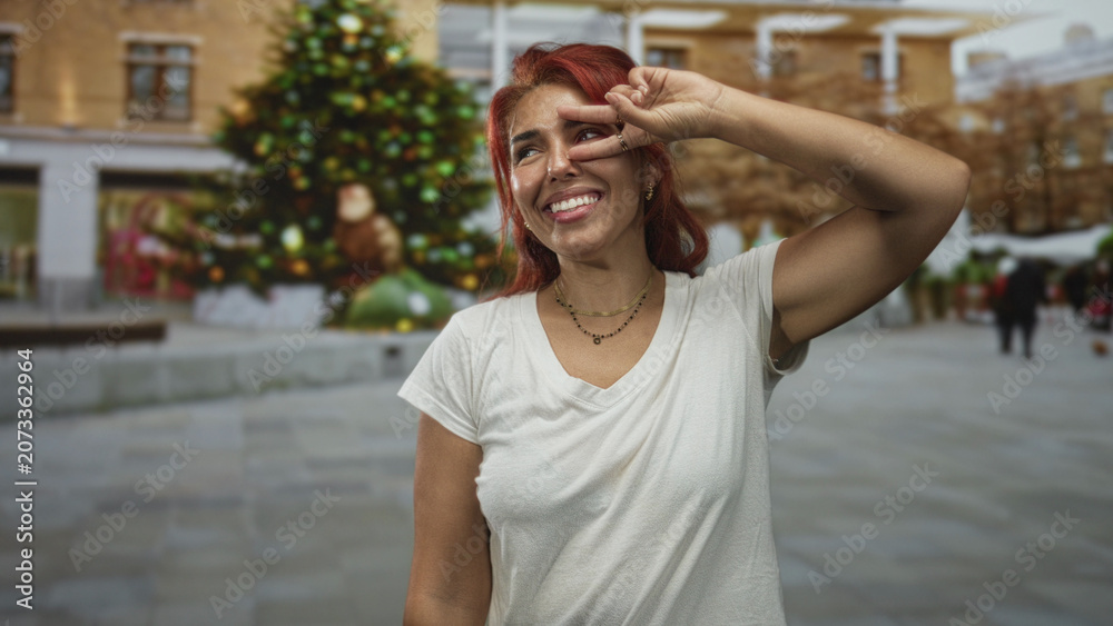 Fototapeta premium Woman showing peace sign with hand and smiling on city street by a large decorated christmas tree; joy playful.