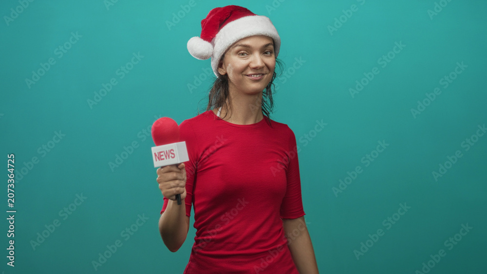 Fototapeta premium Woman wearing santa hat and red shirt holds a news microphone and makes rock hand sign in turquoise studio; festive cheer.