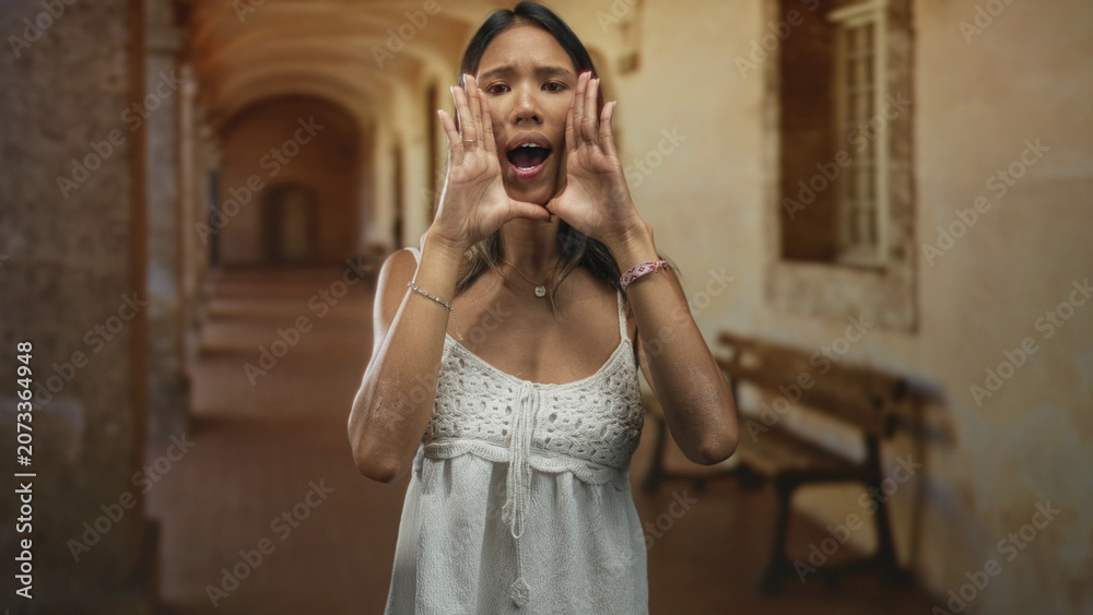 Fototapeta premium Thai woman cupping hands around mouth calling out, showing bare hands and necklace, wearing white crochet dress in a weathered building corridor; urgent appeal.