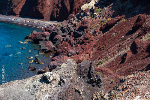 Rugged caldera coastline with crimson lava rocks and clear blue water on Santorini island, Greece.