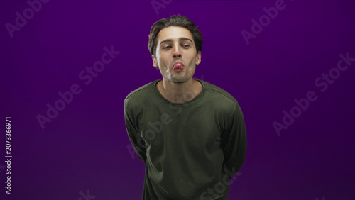 Fotografija Man leaning forward with hands behind back sticks tongue out showing tongue and small hoop earring in studio against purple backdrop; playful mischief