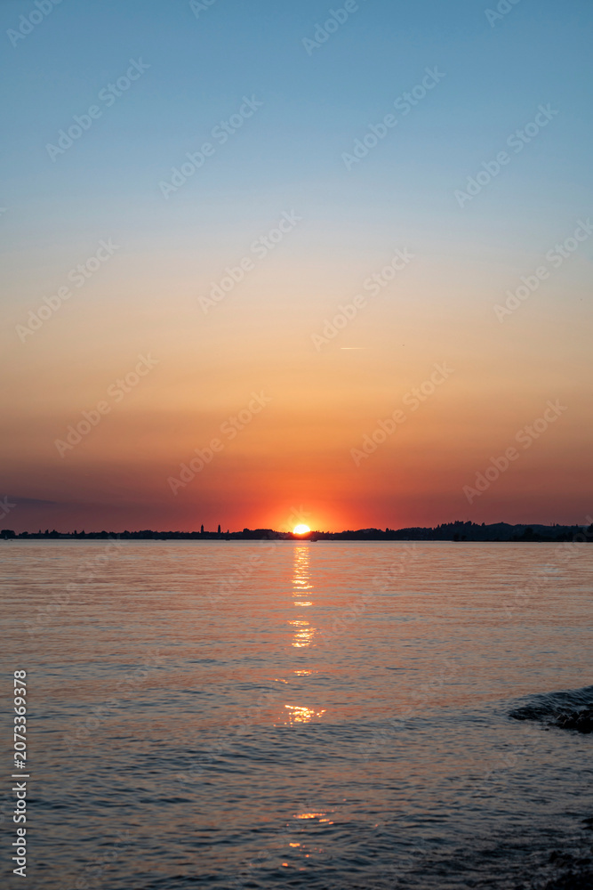 Obraz premium Beautiful sunset over Lake Constance captured from the Austrian shore, showing the vibrant sun setting behind the distant horizon with warm golden reflections on the calm water surface