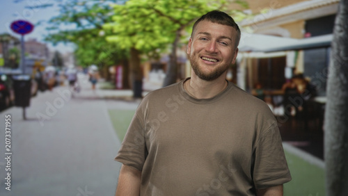 Photos Man smiling with visible forearm and face, relaxed posture, standing near outdoor cafe seating on a busy street; casual contentment