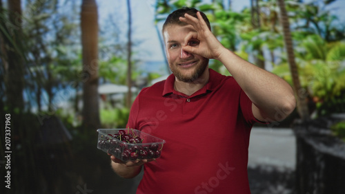 Foto Man holding container of cherries making ok sign with hand near eye in studio; playful summer snack