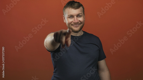 Фотография Man points finger at camera in orange studio showing hand gesture and casual tshirt; playful confidence