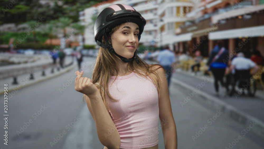 Fototapeta premium Young woman showing pinched fingers gesture with hand on city street, wearing pink tank and helmet; confident attitude.