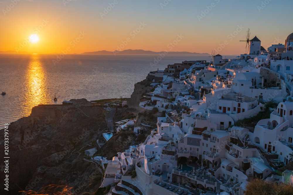 Fototapeta premium Golden sunset reflecting on the Aegean Sea beside whitewashed houses and windmills on the Oia caldera cliffs in Santorini.