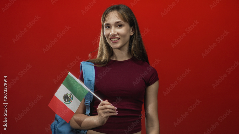 Fototapeta premium Young mexican woman student smiling holding mexican flag and blue backpack in red studio; pride youth optimism.