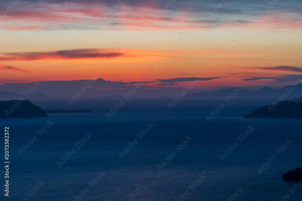 Fototapeta premium Vibrant orange and purple dusk sky above the Aegean Sea and distant island silhouettes, tranquil seascape from Santorini.