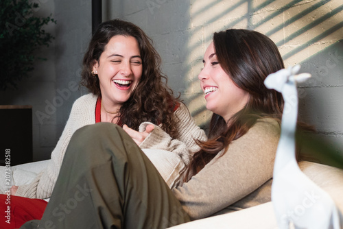 Two young women friends sitting comfortably, sharing a joyful moment, laughing loudly with genuine happiness, and having a casual conversation in a sunlit apartment living room