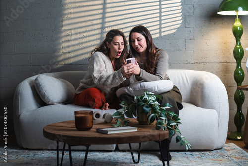 Two young women enjoying leisure time on a comfortable couch, scrolling through exciting content on a mobile phone together while relaxing in a modern living room with sunlight stripes