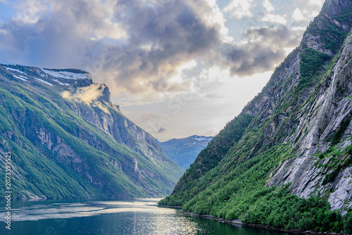 Geiranger, Majestic Norwegian Fjord Landscape, Dramatic fjord landscape with steep, green mountains and calm water under a cloudy sky. Serene natural beauty.