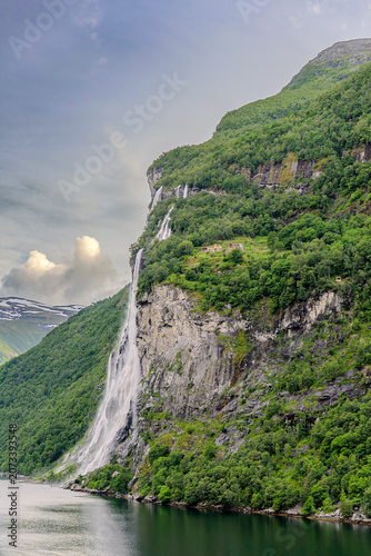 Geiranger, Majestic Norwegian Fjord Landscape, Dramatic fjord landscape with steep, green mountains and calm water under a cloudy sky. Serene natural beauty.