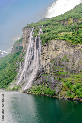 Geiranger, Majestic Norwegian Fjord Landscape, Dramatic fjord landscape with steep, green mountains and calm water under a cloudy sky. Serene natural beauty.