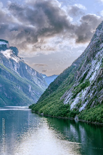 Geiranger, Majestic Norwegian Fjord Landscape, Dramatic fjord landscape with steep, green mountains and calm water under a cloudy sky. Serene natural beauty.