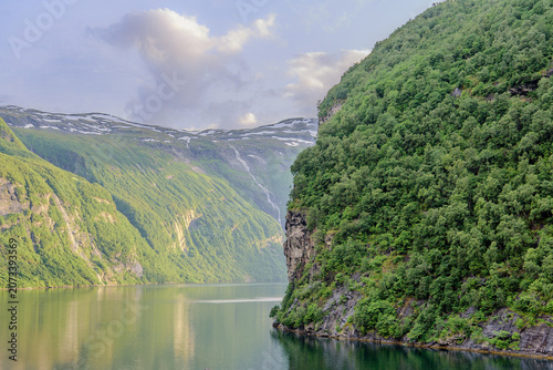 Geiranger, Majestic Norwegian Fjord Landscape, Dramatic fjord landscape with steep, green mountains and calm water under a cloudy sky. Serene natural beauty.