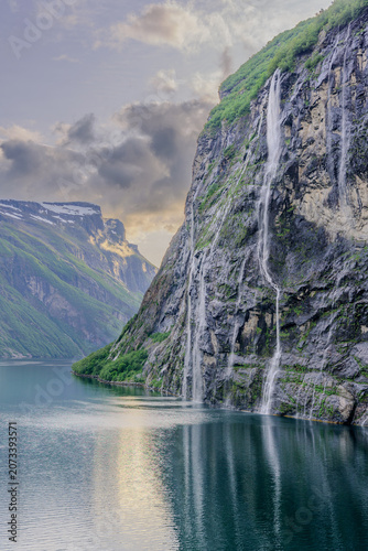 Geiranger, Majestic Norwegian Fjord Landscape, Dramatic fjord landscape with steep, green mountains and calm water under a cloudy sky. Serene natural beauty.