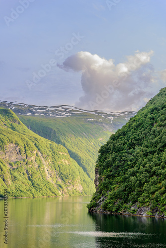 Geiranger, Majestic Norwegian Fjord Landscape, Dramatic fjord landscape with steep, green mountains and calm water under a cloudy sky. Serene natural beauty.