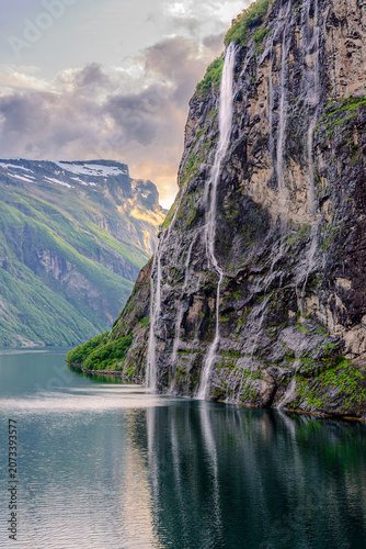 Geiranger, Majestic Norwegian Fjord Landscape, Dramatic fjord landscape with steep, green mountains and calm water under a cloudy sky. Serene natural beauty.