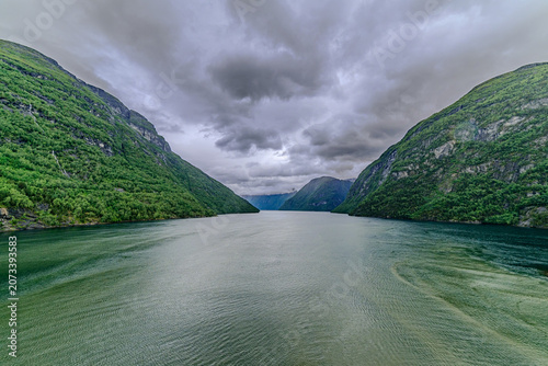 Geiranger, Majestic Norwegian Fjord Landscape, Dramatic fjord landscape with steep, green mountains and calm water under a cloudy sky. Serene natural beauty.