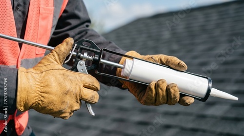 Gloved Caulking Gun Roof Sealant close up worker hand in yellow leather holding metal adhesive tool against dark tiles suitable for diy building supply store posters leak prevention