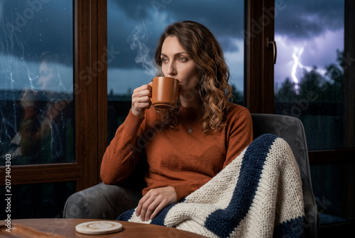 A medium storytelling shot of a woman lifting a dark ceramic mug to her lips, taking a slow sip with realistic wisps of steam rising into the air as lightning flashes outside.
