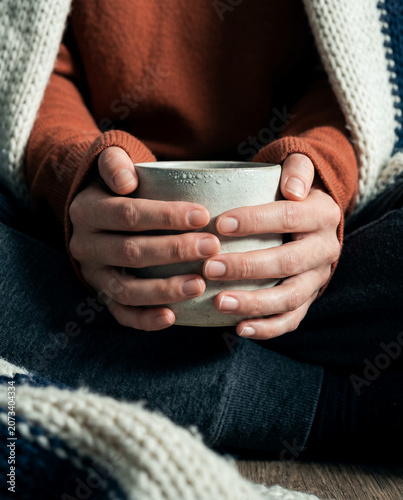 A macro close-up focused on a woman's hands wrapped tightly around a heavy handcrafted ceramic mug with a matte speckled glaze finish under hard side raking light.
