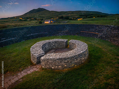 A group of stone blocks forms a circular fortress on the green grass. The mountains rise in the background, bathed in the light of the last moments of sunset. Fields can be seen in the area, along wit