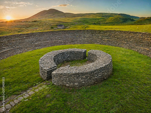 A group of stone blocks creates a circular formation on green grass. Mountains rise in the background under a colorful sky as the sun sets. The area shows fields and a small house nearby.