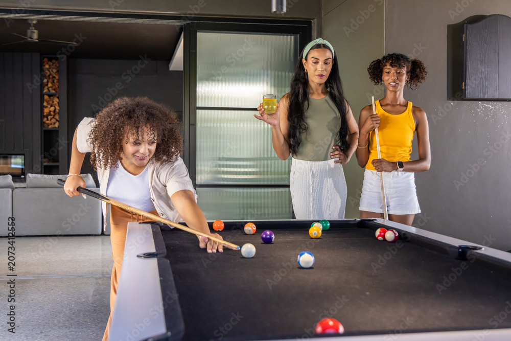 Naklejka premium Diverse female friends playing pool in modern recreation room around black-felt table holding cues