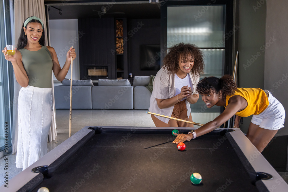 Obraz premium African American female friends leaning over black pool table lining shot with cue in living room